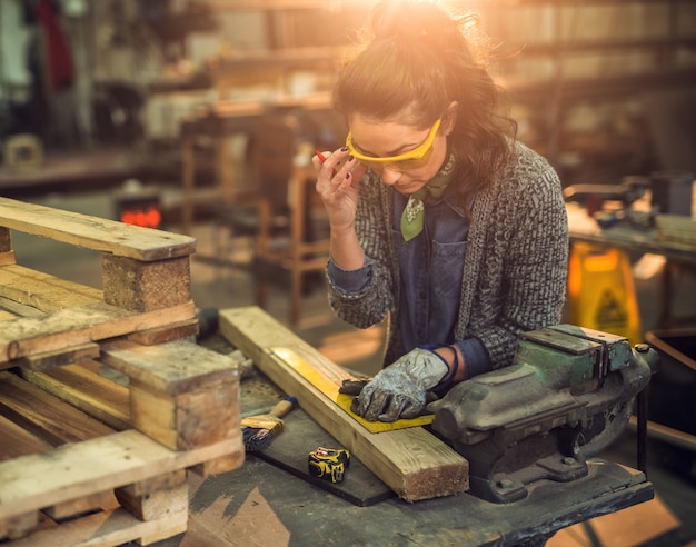 Cerrar vista de trabajadora enfocada profesional serio carpintero mujer trabajando con una regla y haciendo marcas en la madera en la mesa en el taller de tela.
