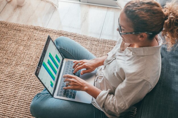 Cerrar la vista trasera de una mujer joven que usa una computadora portátil y una calculadora que trabaja en un proyecto que revisa documentos financieros sentados en la mesa en casa mirando la pantalla de la computadora con diagramas que calculan facturasxA