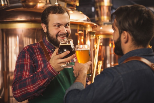 Cervecero alegre barbudo hablando con su asistente, trabajando en la fábrica de producción de cerveza