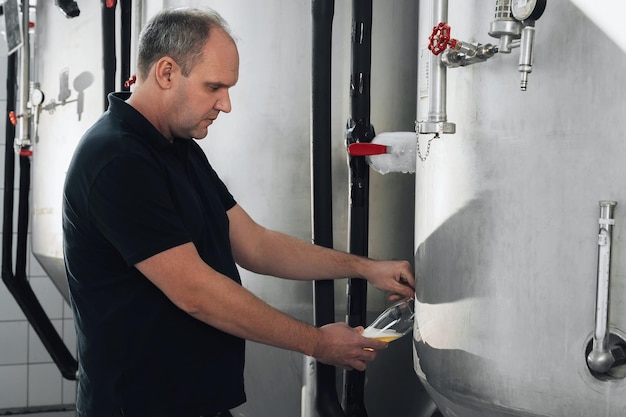 El cervecero en la cervecería vierte cerveza en un vaso del tanque para su inspección