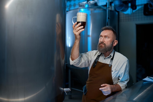 Cervecero senior barbudo que verifica la calidad de la cerveza durante el proceso de tecnología de elaboración de cerveza. Maestro cervecero maduro mirando el nivel de espuma en el vaso