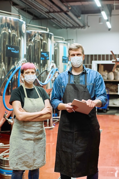 Cerveceros jóvenes en ropa de trabajo y máscaras protectoras de pie frente a la cámara contra enormes tanques de acero para la fermentación y maduración de la cerveza