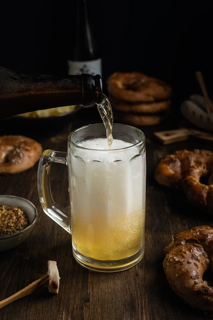 Foto cerveja, derramando em vidro com pretzels, salsicha e salgadinhos na mesa de madeira rústica