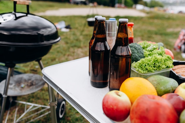 Cerveja, salsichas e legumes frescos na mesa ao ar livre ao lado do churrasco