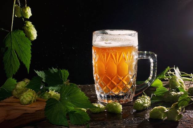 Cerveja sem filtrar bem amarela em uma caneca transparente em um fundo preto com brotos de lúpulo.