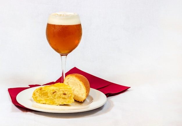 Una cerveza fría servida en una copa de cristal junto con un buen trozo de tortilla española con pan sobre fondo blanco.