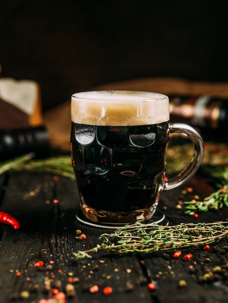 Cerveza oscura en taza con espuma en la mesa de madera con hierbas y pimienta, vertical