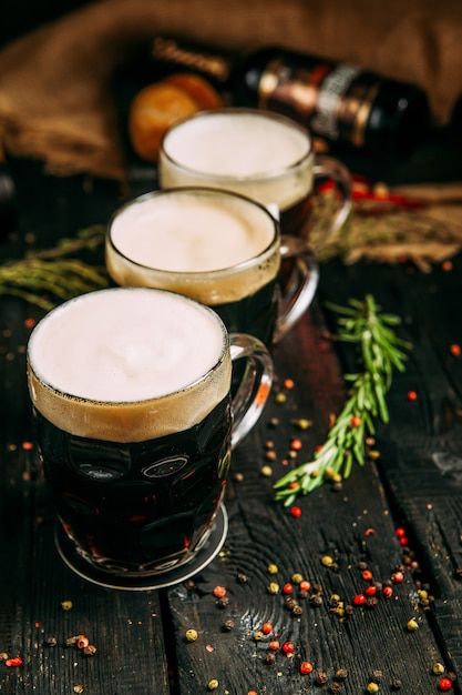 Cerveza oscura en tres tazas con espuma en la mesa de madera con hierbas y pimienta, vertical