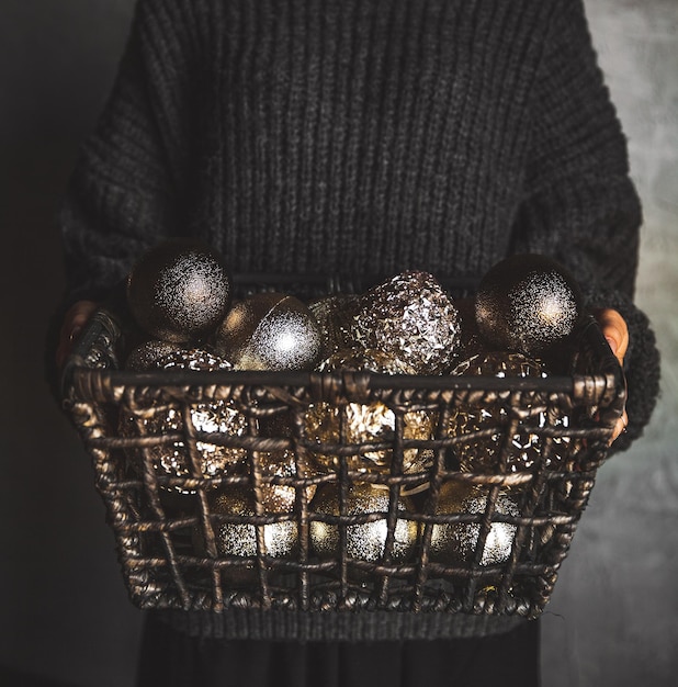 Cesta de arame cheia de brinquedos de decoração vintage dourados de árvore de Natal ou Ano Novo nas mãos de uma senhora em um suéter cinza quente, foco seletivo, corte quadrado