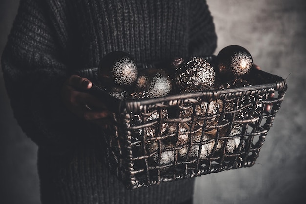 Cesta de arame cheia de brinquedos de decoração vintage dourados de árvore de Natal ou Ano Novo nas mãos de uma senhora em um suéter cinza quente, foco seletivo, corte quadrado