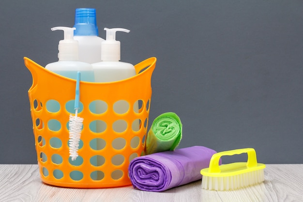 Cesta de plástico com garrafas de detergente, limpador de azulejos, detergente para micro-ondas e fogões. Escova, sacos de lixo em fundo cinza. Conceito de lavagem e limpeza.