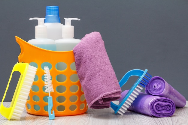 Cesta de plástico com garrafas de detergente, limpador de azulejos, detergente para micro-ondas e fogões. Escovas, toalha, sacos de lixo em fundo cinza. Conceito de lavagem e limpeza.