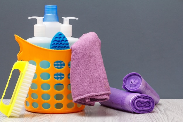 Cesta de plástico laranja com garrafas de detergente, limpador de azulejos, detergente para fornos de micro-ondas e fogões. Escovas, toalha, sacos de lixo em fundo cinza. Conceito de lavagem e limpeza.