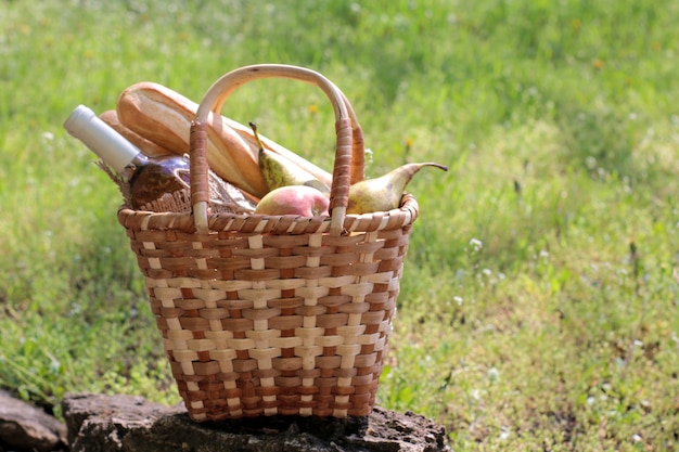 Cesta de vime piquenique com vinho, frutas e outros produtos na grama verde.