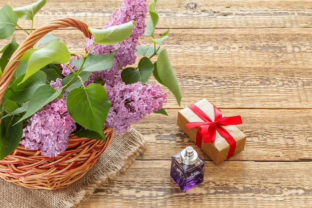 Cesta de mimbre con flores lilas, una botella de perfume y una caja de regalo sobre tablas de madera. Vista superior con espacio de copia.