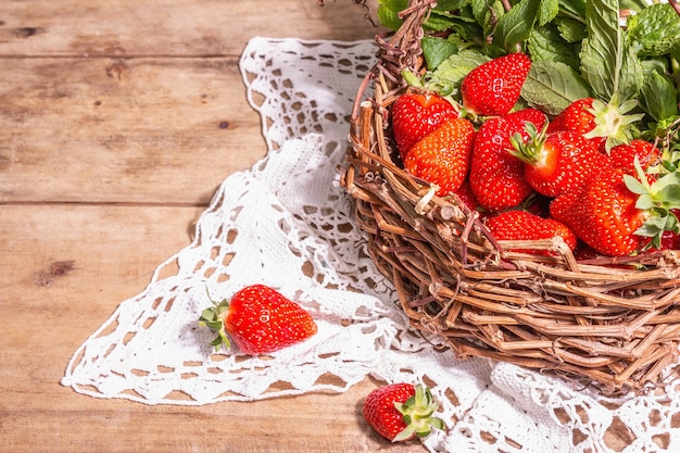 Cesta de mimbre de fresas maduras. Bayas frescas, menta aromática. Mesa de madera antigua, servilleta de ganchillo vintage, estilo rústico, espacio de copia