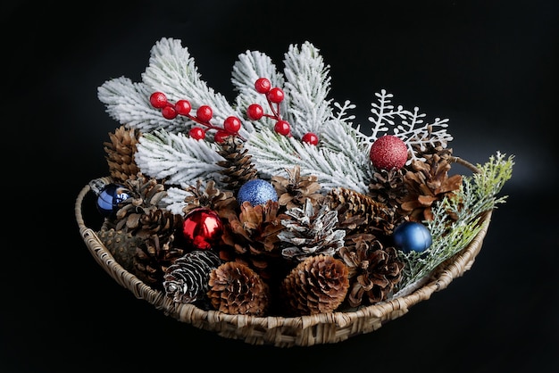 Cesta de Navidad con conos, bolas decorativas de colores y ramas de coníferas cubiertas de nieve sobre un fondo oscuro.