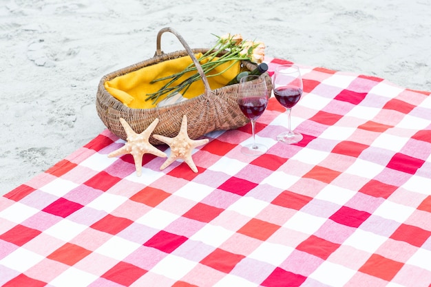 Cesta de picnic con vasos de vino tinto y estrellas de mar sobre una manta en la playa