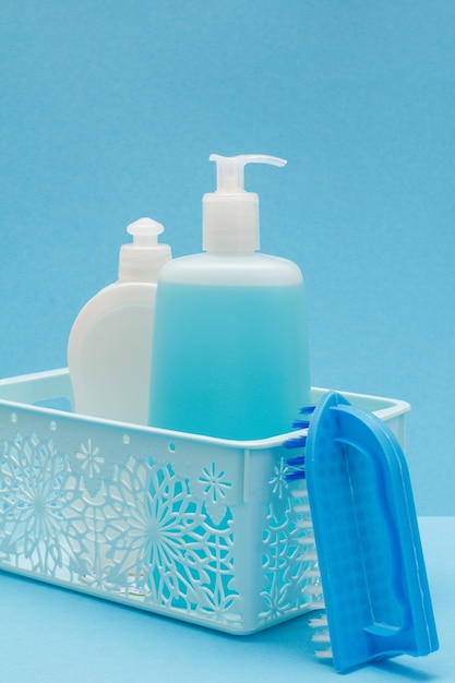 Cesta de plástico con botellas de líquido lavavajillas, limpiador de vidrio y azulejos, cepillo sobre fondo azul. Concepto de lavado y limpieza.