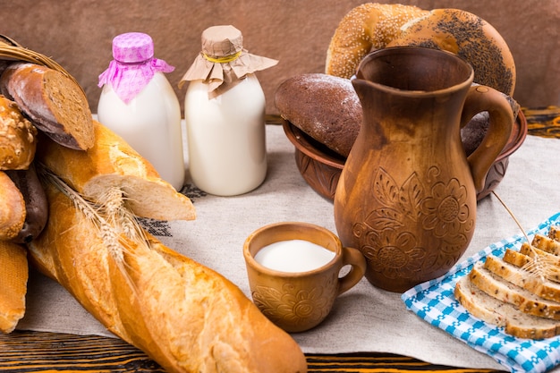 Cesta y tazón de baguettes y bagels junto a una jarra de madera y una taza cerca de dos botellas de leche y una bandeja de pan de molde