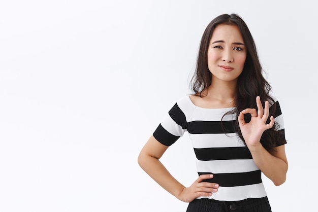 Cética e insegura jovem oriental em camiseta listrada concorda com dúvidas mostra sinal de ok como medida de algo ou julgando aprova a escolha com um sorriso hesitante apertando os olhos em pé fundo branco