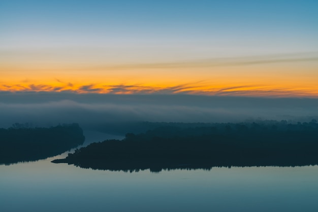 Céu azul adiantado refletido na água do rio. Margem do rio com a floresta sob o céu antes do amanhecer. Faixa amarela no céu pitoresco. Nevoeiro escondeu árvores na ilha. Paisagem atmosférica de mística manhã de natureza majestosa.