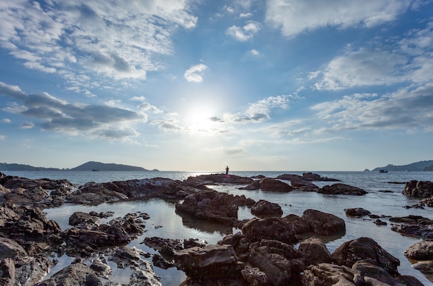 Céu claro nuvens brancas em dia ensolarado e rochas em primeiro plano, homem de pesca silhueta na praia de pedras sob um céu azul de verão.