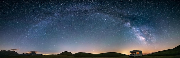 Céu noturno panorâmico sobre as terras altas de campo imperatore, abruzzo, itália. o arco e as estrelas da via láctea sobre a van de campista iluminada. liberdade de campismo na paisagem única das colinas.