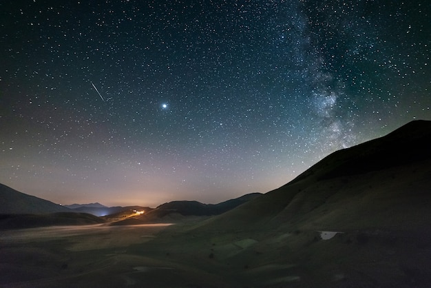 Céu noturno panorâmico sobre Santo Stefano di Sessanio, Abruzzo e Rocca Calascio, Itália. O arco de galáxias da Via Láctea e estrelas sobre a paisagem de colinas únicas de vilarejos iluminados. Planeta Júpiter visível.