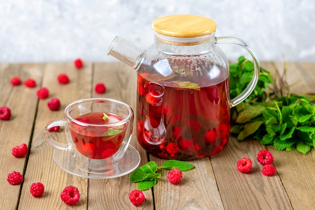 Chá de ervas com frutas vermelhas, framboesas, folhas de hortelã e flores de hibisco em um bule de vidro e xícara na mesa de madeira