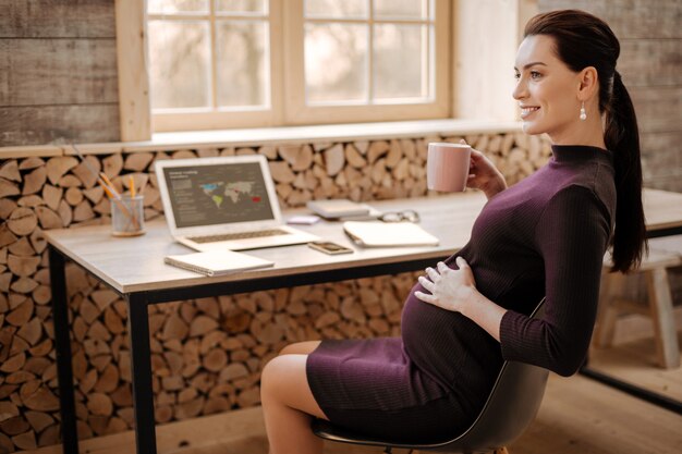 Foto chá de ervas. mulher de negócios grávida otimista e esperançosa acariciando a barriga enquanto sorri e saboreia o chá