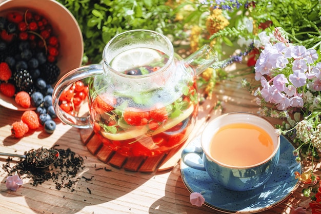 Chá de frutas aromáticas naturais em um bule transparente e em uma xícara entre frutas e flores aquecendo chá aromático com um aroma profundo de frutas e flores silvestres