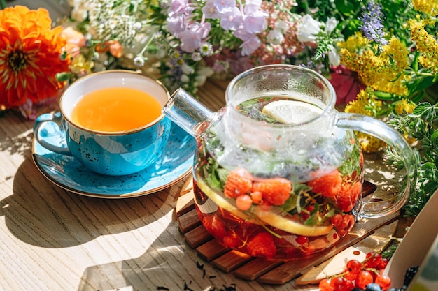 Chá de frutas aromáticas naturais em um bule transparente e em uma xícara entre frutas e flores aquecendo chá aromático com um aroma profundo de frutas e flores silvestres