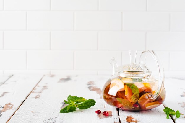 Chá de frutas com folhas de frutas vermelhas, limão, limão e hortelã em um bule de vidro em uma superfície de madeira clara