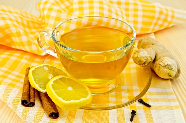 Chá de gengibre em um copo de vidro, duas fatias de limão, canela, cravo, raiz de gengibre em um guardanapo amarelo e uma placa de madeira