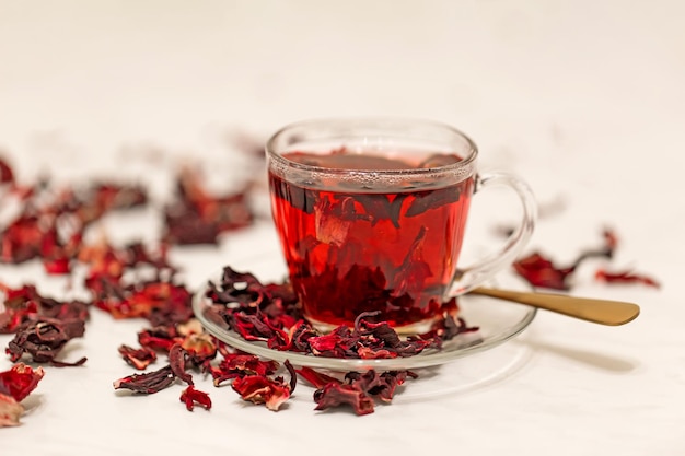 Chá de hibisco vermelho quente em uma caneca de vidro em uma mesa branca entre flores secas da rosa sudanesa