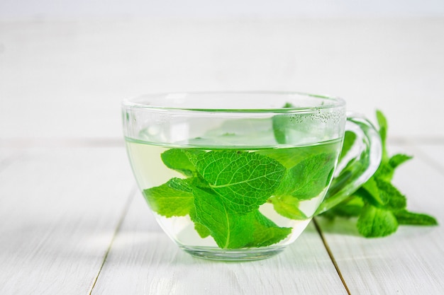 Chá de menta em um copo sobre uma mesa de madeira branca.