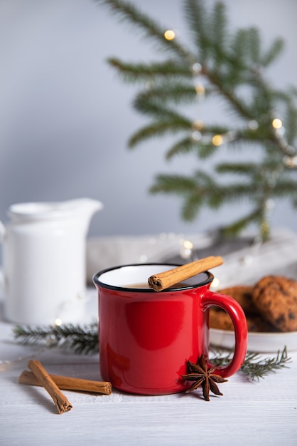 Chá de Natal com canela e especiarias em uma caneca vermelha com biscoitos caseiros em um fundo cinza com uma árvore de Natal e uma guirlanda. Vista frontal