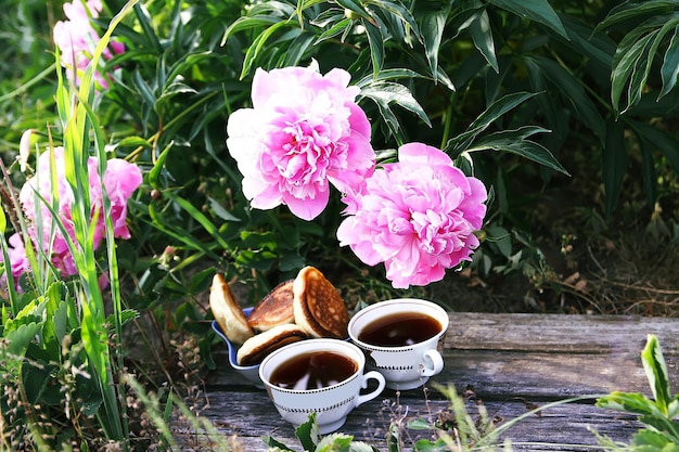 Chá em estilo campestre no jardim de verão na aldeia Duas xícaras de chá preto e panquecas em tábuas de madeira e flores de peônia desabrochando à luz do sol