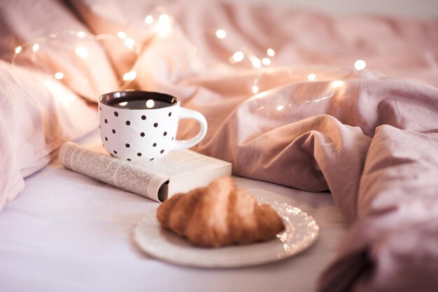 Chá preto fresco na caneca ficar no livro aberto com croissant na cama brilhando luzes closeup. Temporada de férias de inverno. Bom Dia.