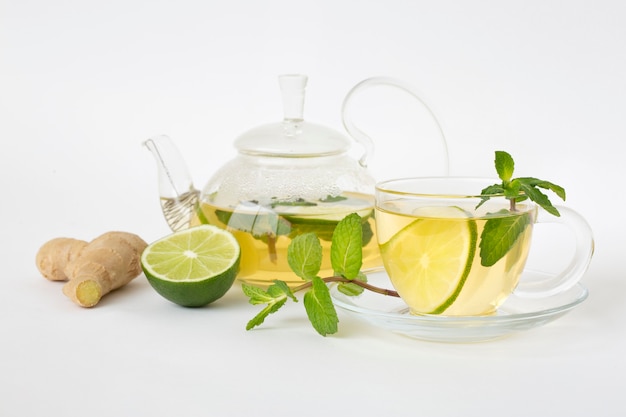 Chá verde em uma xícara de vidro e bule, folhas de hortelã, limão e gengibre em uma mesa branca