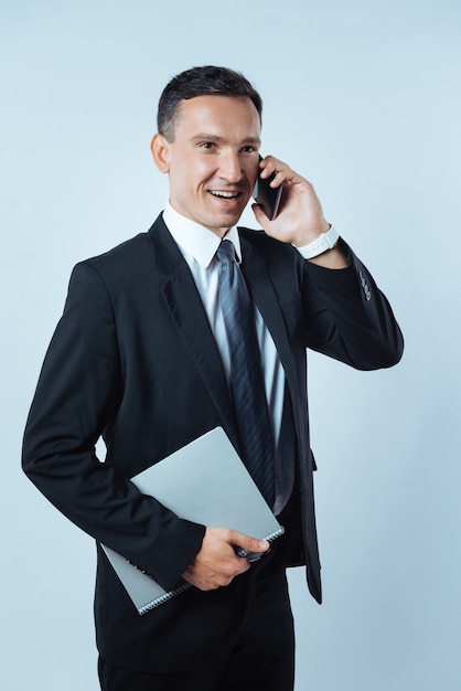 Chamada de negócios. Homem alegre e simpático, encantado, segurando seu telefone celular e fazendo uma ligação enquanto trabalha