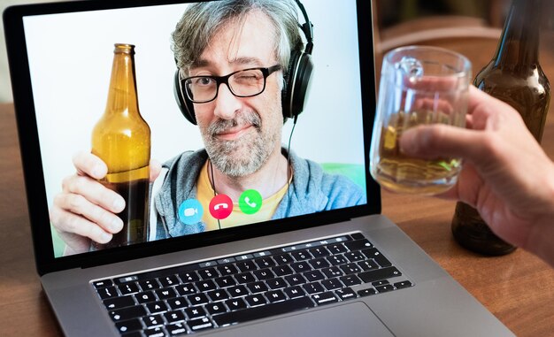 Chamada de vídeo entre amigos, parentes distantes, homem de meia-idade brinda com cerveja junto com outra pessoa usando modernas tecnologias de comunicação