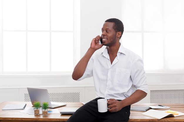 Chamada importante. Jovem empresário negro faz uma ligação no celular enquanto bebe café no interior de um escritório branco moderno, copie o espaço