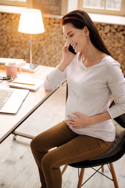 Chamada importante. Vista superior de uma mulher grávida alegre e alegre fazendo uma ligação enquanto ria e acariciava a barriga