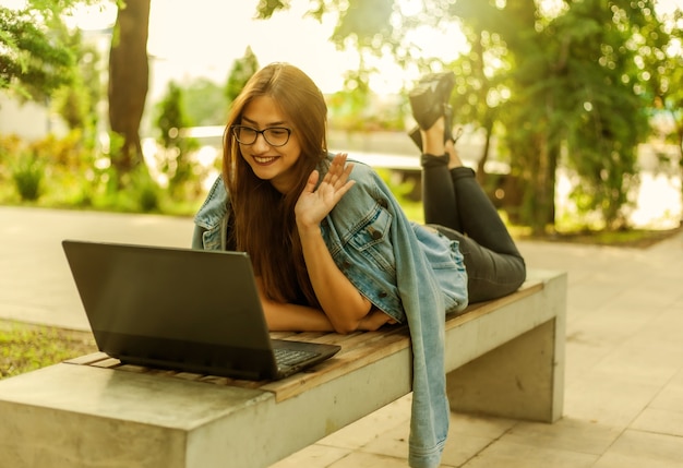 Chamada online. ensino à distância. uma jovem estudante com uma jaqueta jeans e óculos olha para a tela de um laptop enquanto está deitada em um banco em um parque