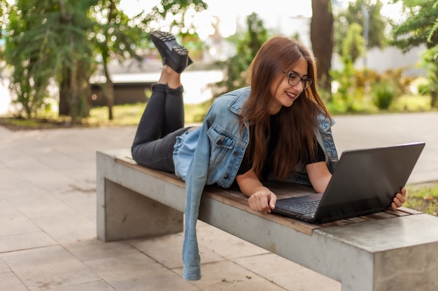 Chamada online. Ensino à distância. Uma jovem estudante com uma jaqueta jeans e óculos olha para a tela de um laptop enquanto está deitada em um banco em um parque