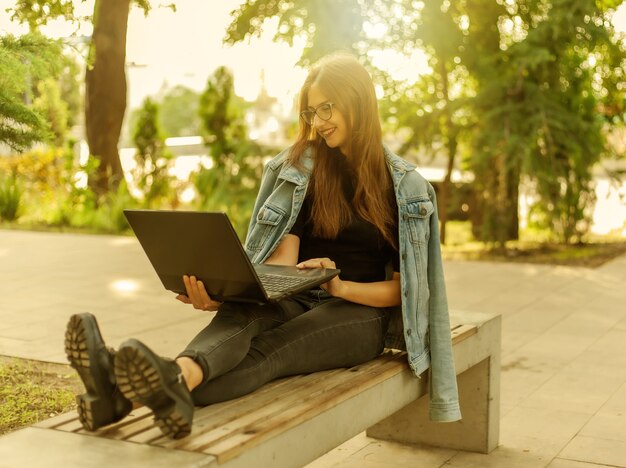 Chamada online. Ensino à distância. Uma jovem estudante com uma jaqueta jeans e óculos olha para a tela de um laptop enquanto está sentada no banco do parque