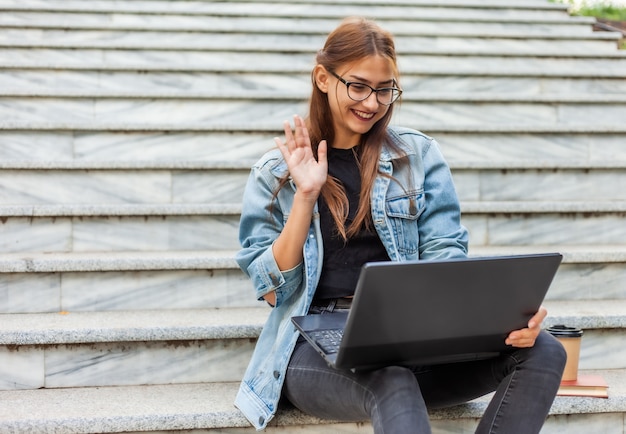 Chamada online. Mulher jovem hippie em jaqueta jeans se comunica com amigos em uma conferência online com laptop enquanto está sentada na escada na cidade