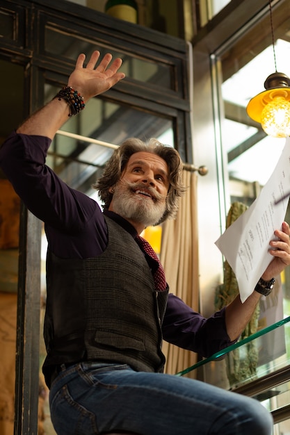 Foto chamando atenção. homem barbudo alegre segurando um menu de café, levantando a mão, chamando um garçom.